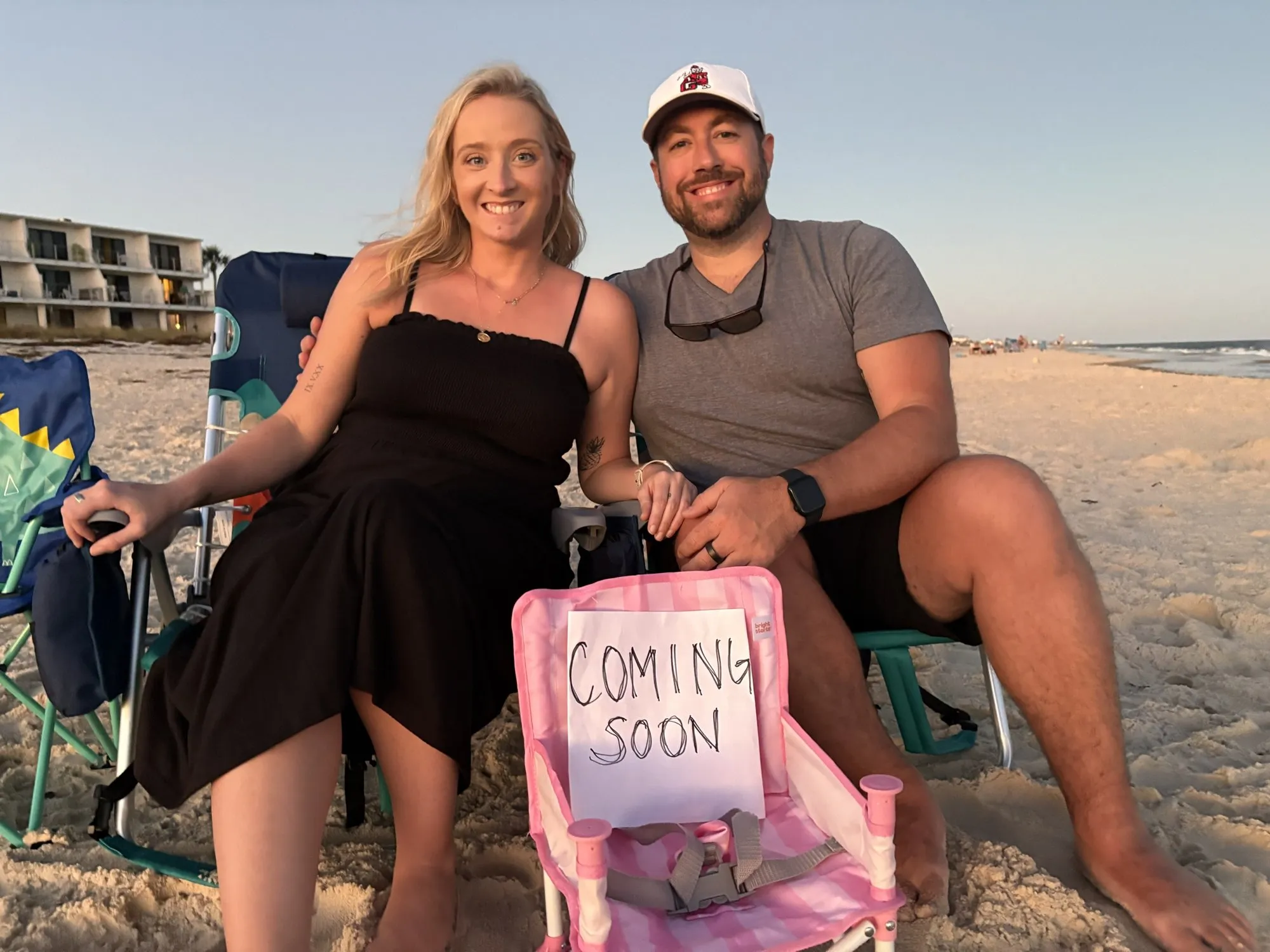 Samantha and Chris on the beach with a pink baby chair and a "Coming soon" sign